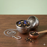 Stainless steel tea infuser ball with chain steeping loose leaf tea in a clear glass cup on a wooden surface with scattered tea leaves.