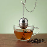 Stainless steel tea infuser ball with chain steeping loose leaf tea in a clear glass cup on a wooden surface with scattered tea leaves.