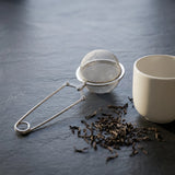 Sleek stainless steel mesh tea strainer with locking clamp beside a ceramic cup and scattered loose leaf tea on dark slate.