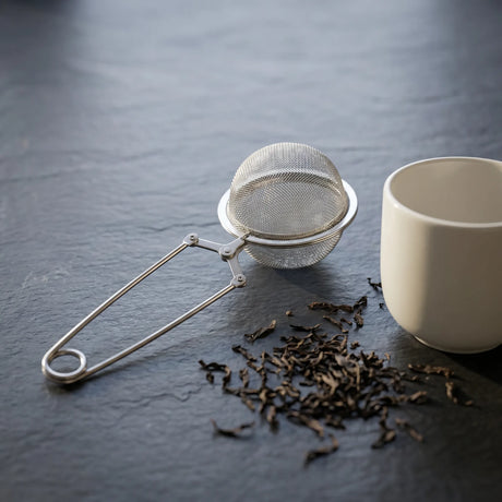 Sleek stainless steel mesh tea strainer with locking clamp beside a ceramic cup and scattered loose leaf tea on dark slate.