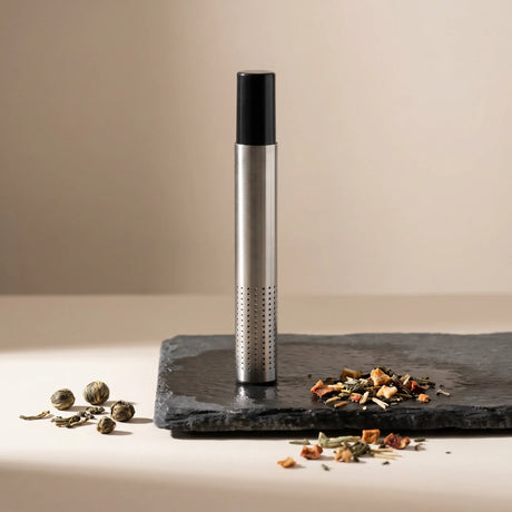 Premium stainless steel tea infuser steeping in a white ceramic mug with steam rising, loose tea leaves scattered on a wooden table