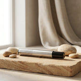 Sleek cylindrical tea strainer with black handle resting on a natural wood board beside smooth river stones